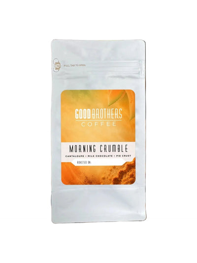 A stand-up pouch of Good Brothers Coffee 'Morning Crumble' coffee stands on a wooden cutting board with flour scattered around it. The package is white with a warm orange and yellow label, and the background shows a softly blurred kitchen scene with a pie and a mug.