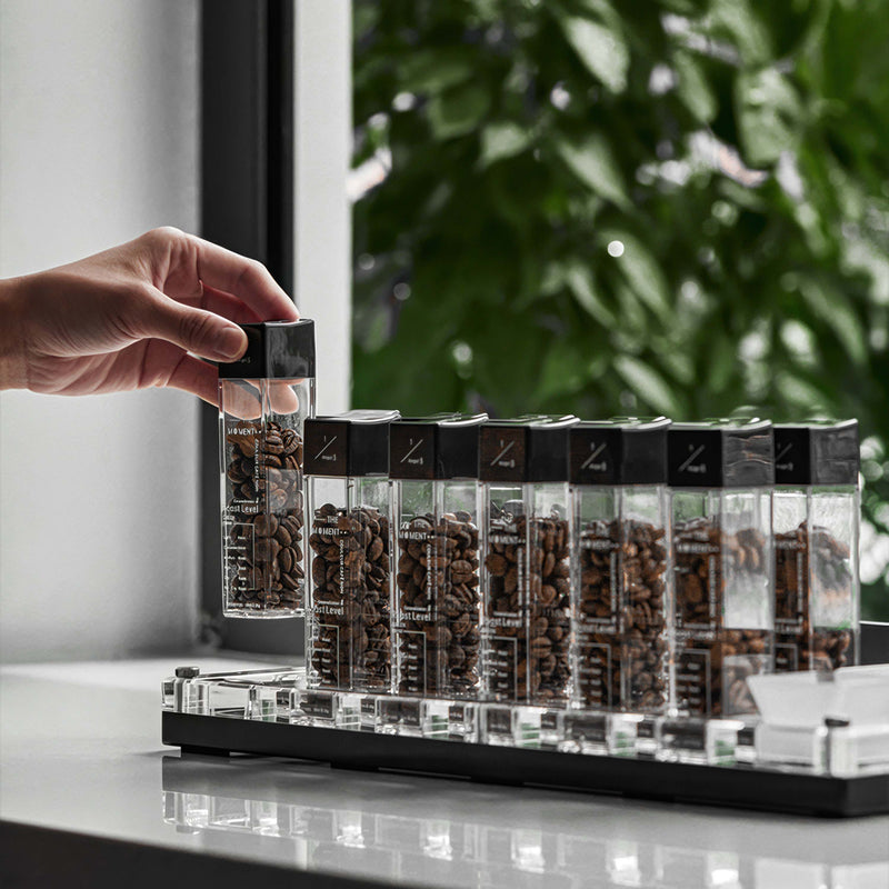 Hand reaching for a container of coffee beans in a transparent holder with black lids.