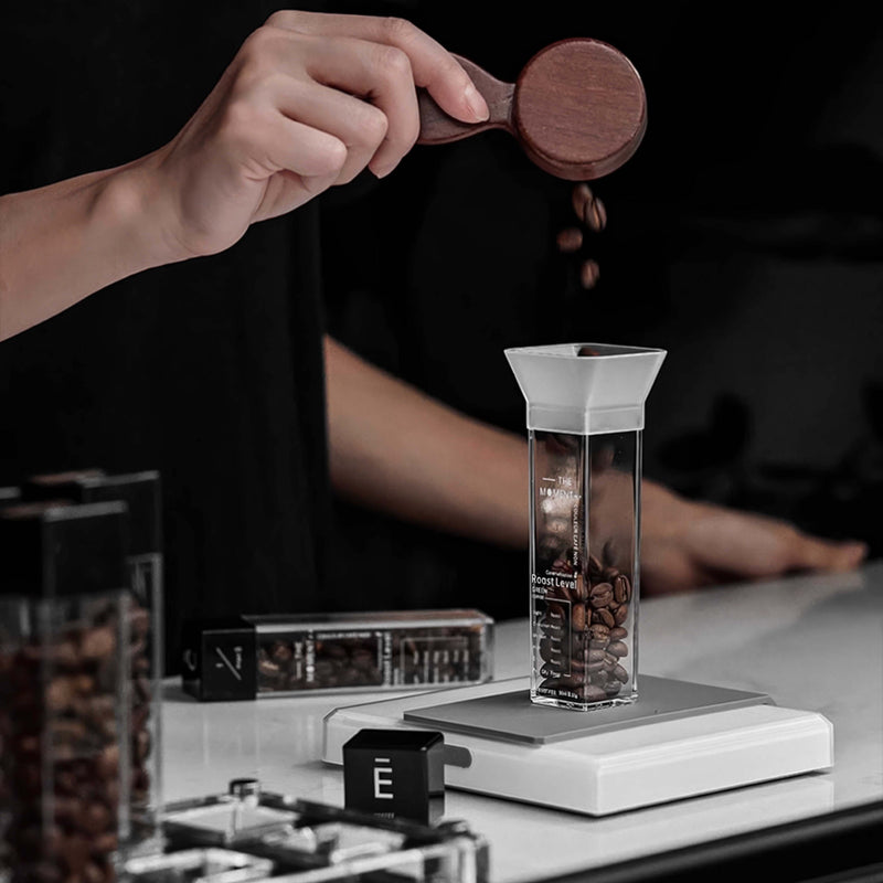 Person using a wooden scoop to measure coffee beans into a transparent container on a dark background